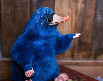 May include: A plush blue creature with a duck-like beak and large eyes. The creature has small hands and feet with black claws. A key and several gold coins are next to the creature. The background is a wooden wall.