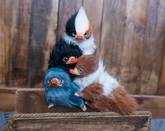 May include: Four plush, bird-like creatures with fluffy fur in various colors: black, blue, brown, and white. Each has a beak, blue eyes, and a tuft of fur on top of their heads. They are stacked on a wooden box.