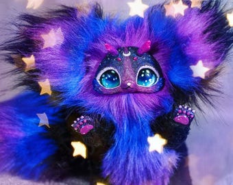 May include: A handmade plush creature with a galaxy theme. The plush features a mix of blue, purple, and black fur, large blue eyes, and star-shaped accents. The creature has small paws and a whimsical design.