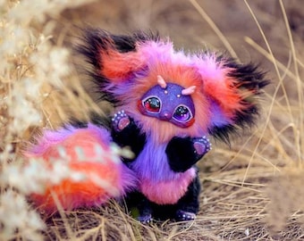 May include: A whimsical, handmade plush creature with a vibrant color palette. The plush features a purple body, pink and orange accents, and large, expressive eyes. Its fluffy tail and fur are a mix of pink, orange, and purple. The creature is set against a natural, outdoor background.