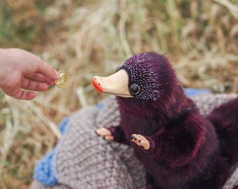 May include: A plush, purple-bodied creature with a cream-colored beak and large, black eyes. The creature is seated on a gray knitted blanket. A hand is offering a gold coin. The background is blurred.
