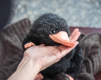 May include: A plush, black creature with a long, orange beak and large, dark eyes. The creature is held in a hand, showcasing its small, detailed feet. The plush has a soft, textured appearance, with white speckles on its head.