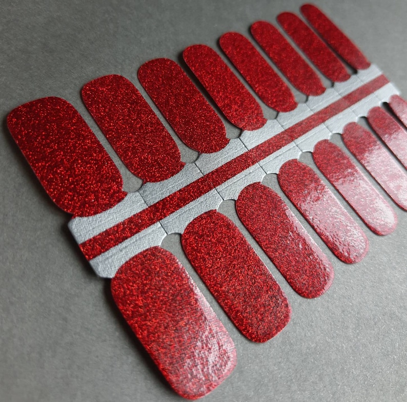 Puede incluir: Tiras de esmalte de u&ntilde;as rojo con purpurina para una manicura festiva. Las tiras est&aacute;n dispuestas en una hoja con un fondo blanco y plateado.