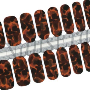 May include: A sheet of nail wraps featuring a tortoiseshell pattern. The wraps have an oval shape and a mix of brown, black, and orange hues. The nail wraps are arranged in rows and are on a silver backing.
