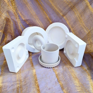 May include: Five white ceramic molds for making various shapes, including a pot, a bowl, and a swirl design. The molds are arranged on a textured, light purple and gold fabric.