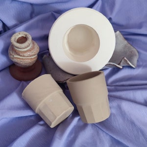 May include: Two beige ceramic cups and a white ceramic planter with a round opening. The cups and planter are on a purple fabric surface.