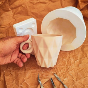 May include: A white ceramic mug with a geometric design. The mug is being held by a hand. There are two small tools and a piece of wood on a brown surface.