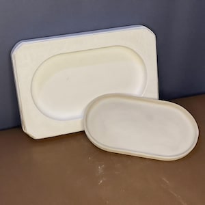 May include: Two off-white, oval-shaped serving trays. One tray is nested inside a larger, rectangular tray with rounded corners. The trays are made of a smooth, matte material and are set against a dark blue and brown background.