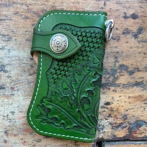 Könnte beinhalten: Ein grüner Leder-Schlüsselanhänger mit einem gewebten Muster und einem Eichel-Design. Der Schlüsselanhänger hat einen silbernen Metallknopf und einen silbernen Metallring.
