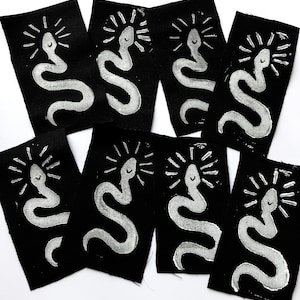 Könnte beinhalten: Acht schwarze Stoffpatches mit weißen gestickten lächelnden Schlangen mit einem Sonnenstrahl-Design darüber.