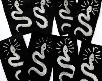 Op de afbeelding: Acht zwarte stoffen patches met witte geborduurde glimlachende slangen met een zonnestralenontwerp erboven.