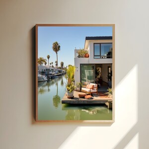 Op de afbeelding: Een ingelijste wandkunstprint met een modern wit huis met een houten terras boven een rustig kanaal. De sc&egrave;ne toont een terrasbank met oranje kussens, potplanten en palmbomen onder een heldere blauwe lucht die reflecteert in het water.