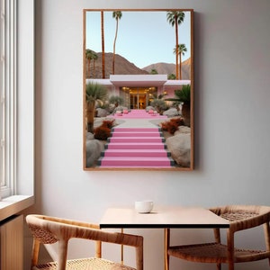 May include: A framed wall art print depicting a modern house with bright pink stairs leading to the entrance. The scene features desert landscaping with palm trees, large rocks, and brown shrubs set against a backdrop of mountains under a clear sky.