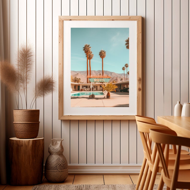 Op de afbeelding: Een ingelijste kunstprint met een woestijnmotel, een zwembad, palmbomen en bergen onder een heldere lucht. Het bord toont de tekst MOTEL in dikke letters. De print zit in een lichthouten lijst en hangt aan een witte wand met verticale panelen.