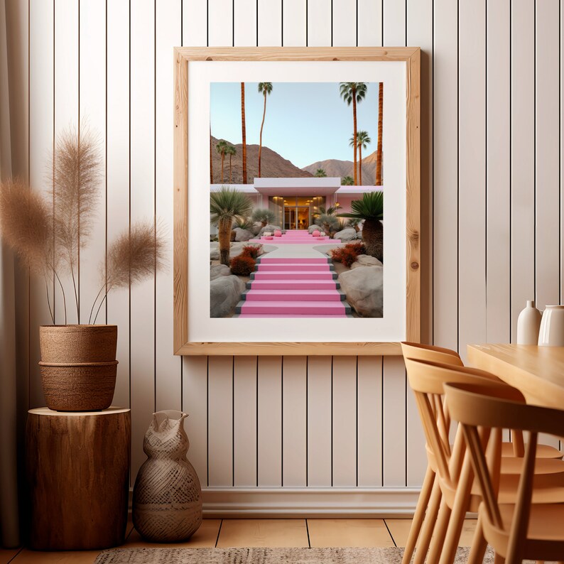 May include: A framed wall art print featuring a desert landscape with a modern building and a set of bright pink stairs leading to the entrance, surrounded by large rocks, palm trees, and a mountain range under a clear blue sky.