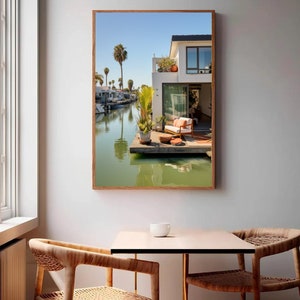 Op de afbeelding: Een ingelijste wandkunstprint met een sc&egrave;ne aan het water met een houten terras, een wit huis, palmbomen en een kanaal. Het terras is voorzien van een stoel met oranje kussens, potplanten en een kleine tafel onder een helderblauwe lucht.