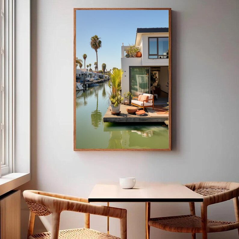 Op de afbeelding: Een ingelijste wandkunstprint met een sc&egrave;ne aan het water met een houten terras, een wit huis, palmbomen en een kanaal. Het terras is voorzien van een stoel met oranje kussens, potplanten en een kleine tafel onder een helderblauwe lucht.