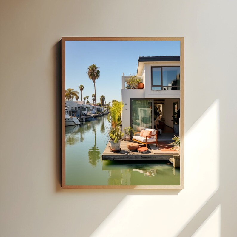 Op de afbeelding: Een ingelijste wandkunstprint met een modern wit huis met een houten terras boven een rustig kanaal. De sc&egrave;ne toont een terrasbank met oranje kussens, potplanten en palmbomen onder een heldere blauwe lucht die reflecteert in het water.