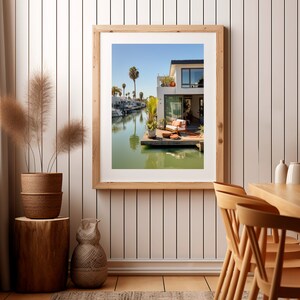 Op de afbeelding: Een ingelijste kunstprint van een huis aan het water met een houten terras, wit buitenmeubilair en potplanten. De sc&egrave;ne toont een kanaal met boten, palmbomen en een helderblauwe lucht, gepresenteerd in een lichthouten lijst aan de muur.