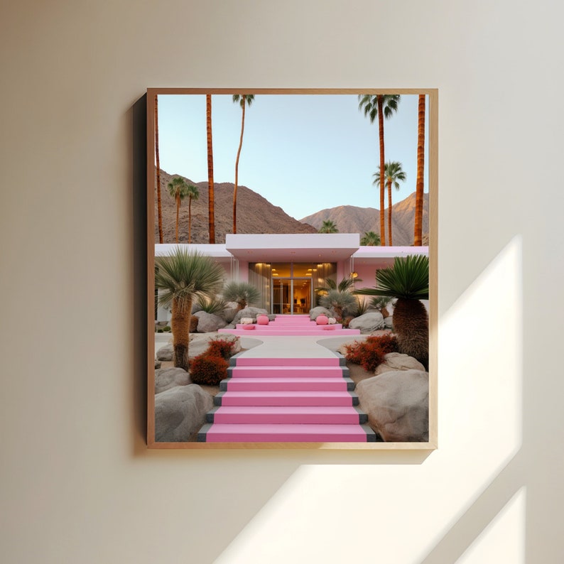 May include: A framed wall art print depicting a modern house with bright pink stairs leading to the entrance. The scene features desert landscaping with palm trees, large grey boulders, and reddish-brown shrubs against a backdrop of mountains.