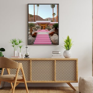 May include: A framed wall art print depicting a desert landscape with a modern building featuring a bright pink staircase leading to the entrance, surrounded by palm trees, large rocks, and a clear blue sky in a sunny, arid environment.