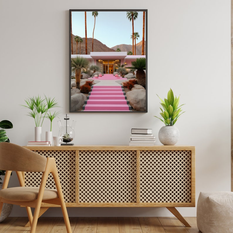 May include: A framed wall art print depicting a desert landscape with a modern building featuring a bright pink staircase leading to the entrance, surrounded by palm trees, large rocks, and a clear blue sky in a sunny, arid environment.
