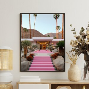 May include: A framed wall art print depicting a modern house with bright pink stairs leading to the entrance. The scene features tall palm trees, desert landscaping with large rocks, and mountains in the background under a clear sky.
