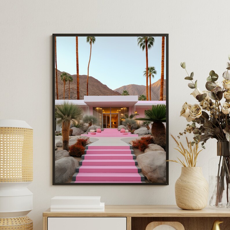 May include: A framed wall art print depicting a modern house with bright pink stairs leading to the entrance. The scene features tall palm trees, desert landscaping with large rocks, and mountains in the background under a clear sky.