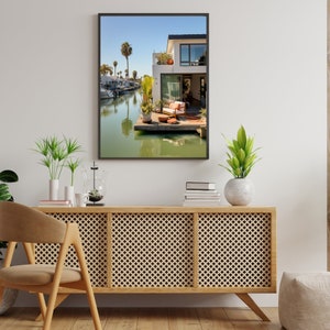 Op de afbeelding: Een ingelijste kunstdruk van een modern wit huis met een houten terras aan een kanaal, met palmbomen, een ligstoel en potplanten onder een helderblauwe lucht, weergegeven boven een houten dressoir met een latwerkpatroon.