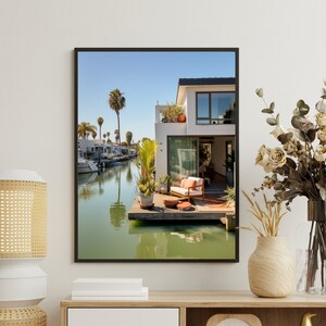 Op de afbeelding: Een ingelijste muurkunstprint met een modern wit huis aan een kanaal met een houten terras, een witte bank met oranje kussens, potplanten en palmbomen onder een helderblauwe lucht, tentoongesteld in een kamer met een lamp en een vaas bloemen.