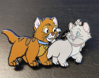 May include: A pin featuring two cartoon cats. The cat on the left is orange and white with a blue collar and a bell. The cat on the right is white with a pink bow and a pink collar.