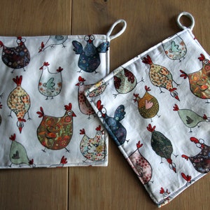 Op de afbeelding: Twee witte katoenen ovenwanten met een kleurrijk kippenpatroon. De kippen zijn geïllustreerd in een grillige stijl met verschillende kleuren en patronen.