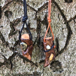 Könnte beinhalten: Zwei Holzanhänger mit Lederbändern. Die Anhänger sind aus kleinen Holzstücken mit natürlicher Rinde gefertigt und haben kleine Steine eingebettet. Ein Anhänger hat ein schwarzes Band und der andere ein braunes Band.
