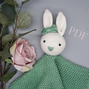 Könnte beinhalten: Ein gehäkeltes Hasen-Kuscheltier mit einer grünen Decke. Der Hase hat ein weißes Gesicht mit schwarzen Augen und einer grünen Nase. Der Hase trägt eine grüne Schleife.