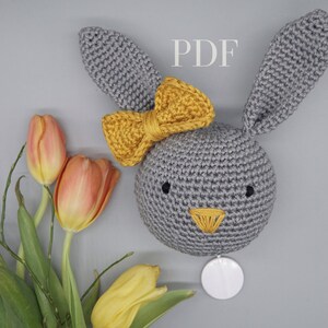 Könnte beinhalten: Ein grauer gehäkelter Hasenkopf mit einer gelben Schleife. Der Hase hat schwarze Augen und eine gelbe Nase. Der Hase sitzt auf einer grauen Oberfläche mit einem weißen Kreis dahinter.
