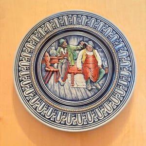 May include: A round ceramic decorative plate featuring a relief scene of two figures in a tavern setting. One figure stands in a green jacket, while the other sits on a bench wearing a red apron. The border has a blue and grey floral pattern.