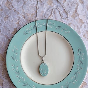May include: A silver-toned necklace featuring an oval pendant with a light blue background and a delicate white floral pattern. The pendant hangs from a thin silver chain, matching the floral design on the light blue and white plate.