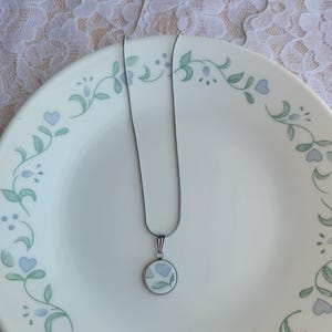 May include: A silver-toned necklace with a round pendant featuring a blue floral design. The pendant is set against a white background. The necklace rests on a white plate with a blue and green floral border.