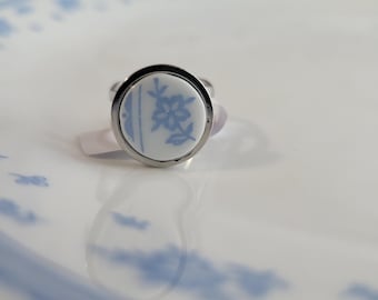 May include: A silver-toned ring with a round, white face featuring a blue floral design. The ring is set against a white plate with blue floral patterns. The ring's design is reminiscent of vintage porcelain.