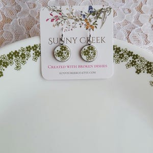 Puede incluir: Pendientes redondos con fondo blanco y estampado floral verde. Los pendientes están sobre una tarjeta blanca con el texto "SUNNY CREEK" y "CREATED WITH BROKEN DISHES". Los pendientes se exhiben en un plato blanco con un estampado floral verde.