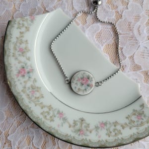 May include: A silver adjustable bracelet with a round charm featuring a floral design of pink roses. The bracelet is displayed on a white plate with a decorative floral border.