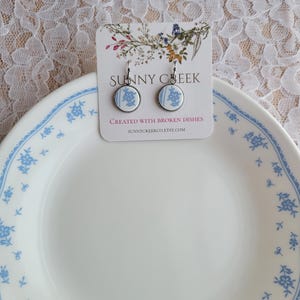 Puede incluir: Pendientes redondos con diseño floral azul sobre fondo blanco. Los pendientes se muestran en una tarjeta con el texto "SUNNY CREEK" y "CREATED WITH BROKEN DISHES". Se colocan sobre un plato blanco con detalles florales azules.