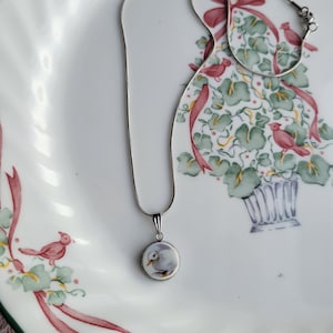 May include: A silver-toned snake chain necklace featuring a small, circular pendant. The pendant displays a miniature illustration of a white duck with a yellow beak against a light background, encased in a simple metal setting.