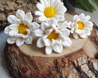 May include: Four white daisy flowers with yellow centers are arranged on a wooden slice. The flowers are made of polymer clay and are perfect for crafting.