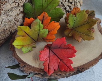 Puede incluir: Cinco hojas de arcilla polimérica hechas a mano en tonos de verde, naranja y rojo. Las hojas están dispuestas sobre una rodaja de madera.