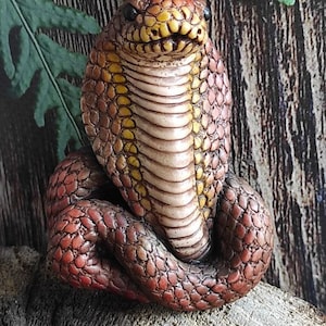 May include: A small, hand-painted ceramic cobra figurine in a red, brown and yellow colour scheme. The cobra is coiled with its head raised and its hood expanded.