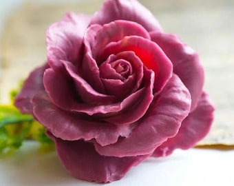 May include: A sculpted rose flower in a deep mauve-purple color with layered, curved petals that spiral toward the center. The surface has a smooth, matte texture, and the flower is shown from a top-down angle with a small green leaf visible.