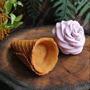 May include: A light brown waffle cone sits next to a pink rose-shaped confection on a wooden surface.