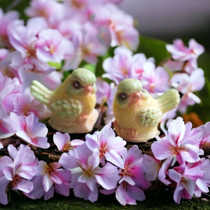 Può includere: Due piccole statuine di uccelli in ceramica dipinte di verde e giallo, posate su un letto di fiori rosa.
