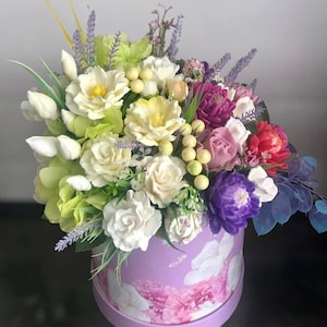 Puede incluir: Una caja morada con un patrón floral contiene un ramo de flores artificiales. El ramo incluye rosas blancas, rosas amarillas, tulipanes blancos, lavanda y flores moradas y rojas.